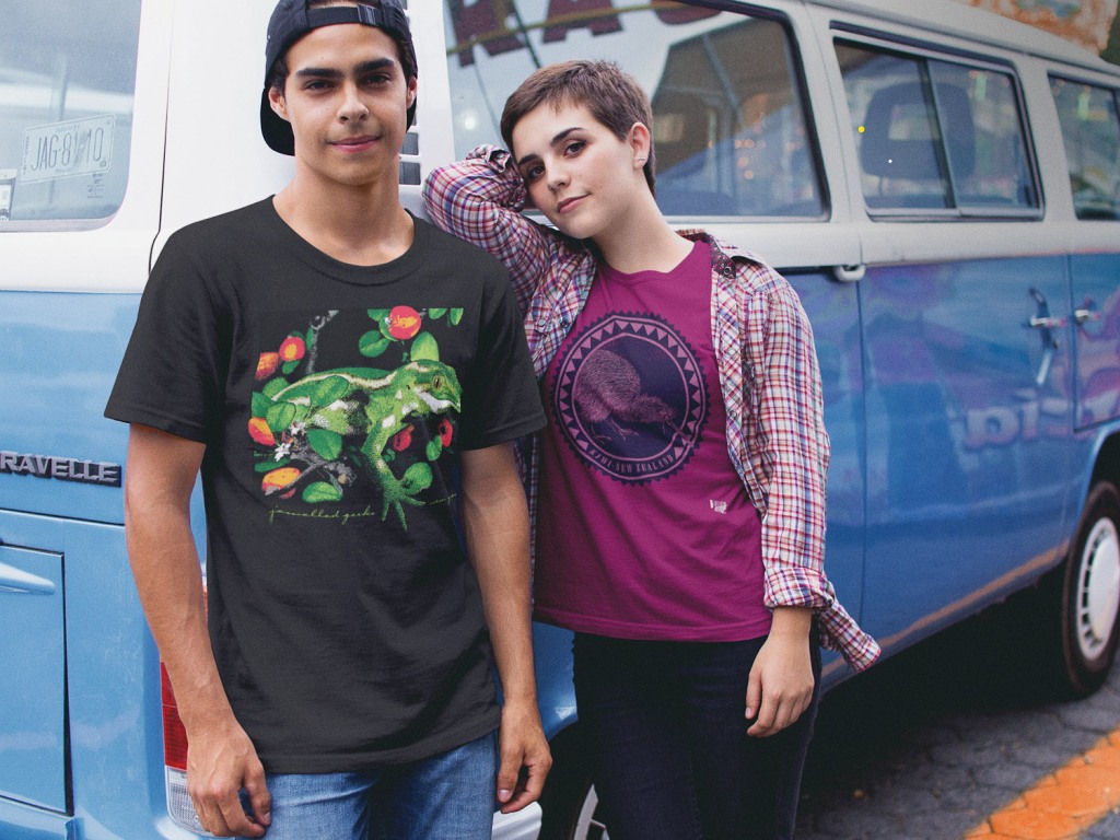 A young couple wearing two different Surface Active New Zealand nature t-shirts leaning against their VW kombi campervan. He is wearing the jewelled gecko design on a black t-shirt, she is wearing a cardinal red iconic kiwi roundel t-shirt.