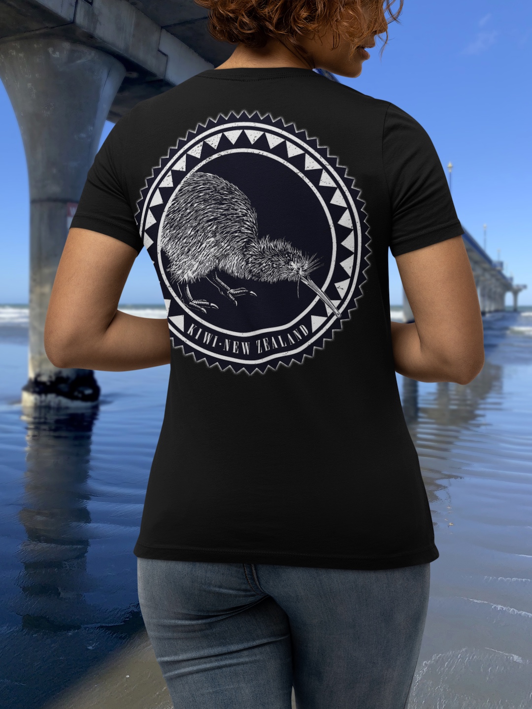 Back view of a curly-haired woman modelling the the large art of a black Surface Active iconic Kiwi, New Zealand t-shirt beside the New Brighton pier.